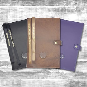 May include: Three leather-bound journals with gold lettering and a clear window on the front cover. The journals are brown, black, and purple. The text on the journals reads "Living the Steps" and "One Day At a Time".