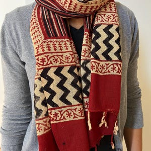 May include: A red, black, and cream cotton scarf with a geometric pattern. The scarf has a fringe on the ends.