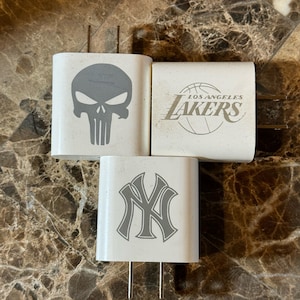 May include: Three white phone chargers with different designs. The first charger has a gray skull and crossbones design. The second charger has a gold basketball and the text "LOS ANGELES LAKERS". The third charger has a gray New York Yankees logo.