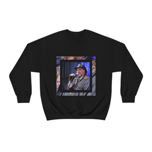 May include: Black sweatshirt with a photo of a man wearing a cowboy hat and singing into a microphone. The photo is framed with a white border.