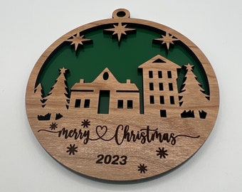 May include: A round wooden Christmas ornament with a green background and a cutout design of houses, trees, and stars. The ornament is inscribed with "merry heart Christmas" and "2023".