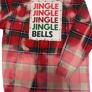 Może przedstawiać: Czerwono-zielona koszula w kratę z efektem wybielania. Biała naszywka na plecach zawiera słowa "JINGLE JINGLE JINGLE JINGLE BELLS" w czerwonych, zielonych i czarnych literach. Naszywka jest obrysowana małymi złotymi ćwiekami.