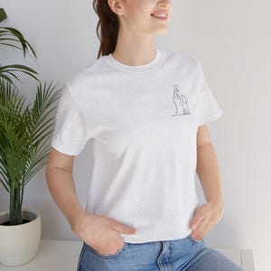 Könnte beinhalten: Ein hellgraues Kurzarm-T-Shirt mit einer kleinen schwarzen Strichzeichnung auf der linken Brust. Das Motiv zeigt eine Hand, die ein Fingerherz über einem kleinen Hund formt, wobei ein winziges Herz über den Fingern schwebt.