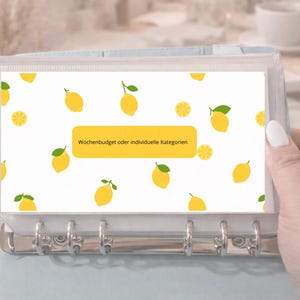 Könnte beinhalten: Ein klarer Ordner mit einem weißen Blatt mit einem Zitronenmuster. Das Blatt hat ein gelbes Rechteck mit dem Text "Wochenbudget oder individuelle Kategorien". Der Ordner wird von einer Hand gehalten.