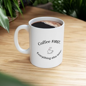 May include: A white ceramic mug filled with dark coffee, featuring the black printed text Coffee FIRST. Everything else later. and a small line drawing of a coffee cup with a heart-shaped steam swirl centered between the two lines of text.