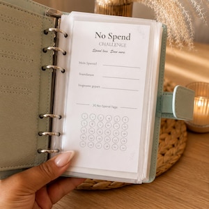 Könnte beinhalten: Ein hellgrüner Ringbuchplaner mit sechs Ringen und einer transparenten Kunststoffhülle für eine No-Spend-Challenge. Die Seite enthält den Text No Spend CHALLENGE, Spend less. Save more., Mein Sparziel, Startdatum, Insgesamt gepart und 30 No-Spend-Tage.
