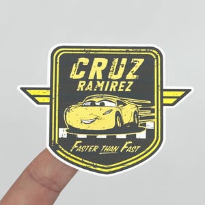 May include: A yellow and black Cruz Ramirez sticker with a cartoon car graphic. The sticker has the text "CRUZ RAMIREZ" and "Faster Than Fast". The sticker is in a shield shape with wings.