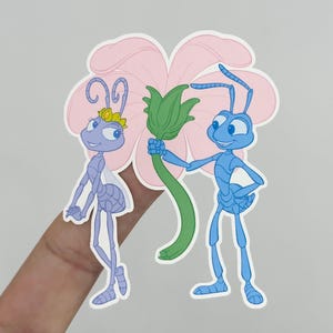 May include: A sticker featuring two cartoon ants. One is light purple with a yellow crown, and the other is blue, holding a green stem with a pink flower in the background. The sticker is held by a hand.