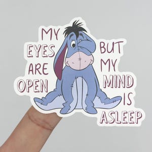 May include: A sticker featuring Eeyore from Winnie the Pooh, in shades of blue, gray, and pink. The text reads "MY EYES ARE OPEN BUT MY MIND IS ASLEEP" in a maroon font. The sticker is held by a finger.
