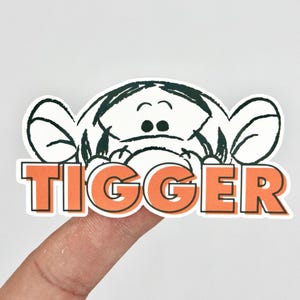 May include: A sticker featuring the cartoon character Tigger from Winnie the Pooh. The sticker shows Tigger's face in black and white, with the name "TIGGER" in orange block letters below.