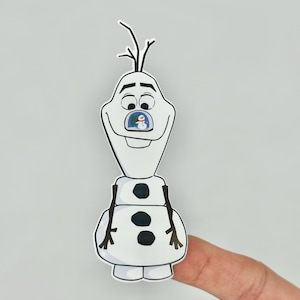 May include: A white Olaf sticker with black accents, featuring a smiling face and twig hair. The sticker has a small image of a snowman inside Olaf's mouth. The sticker is held by a finger.