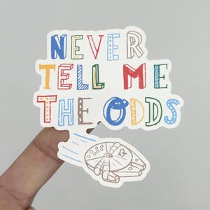 May include: A colorful sticker with the text "NEVER TELL ME THE ODDS" in various colors. Below the text is a drawing of the Millennium Falcon spaceship, with a blue trail behind it.