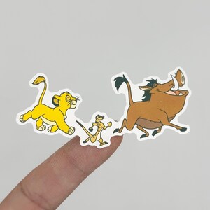 May include: A sticker set featuring three characters from the animated film, The Lion King. The characters include a yellow lion cub, a meerkat, and a brown warthog, all in a running pose. The stickers are on a white background.