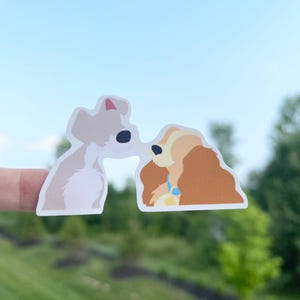 May include: A sticker featuring two cartoon dogs kissing. One dog is light tan with a pink ear, and the other is brown with long hair and a blue collar. The background is a bright blue sky and green grass.