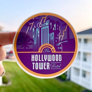 May include: A round sticker featuring a purple and orange design with the text "The Hollywood Tower Hotel." The sticker depicts a stylized image of a building with lightning bolts and palm trees. The background shows a green lawn and a building.