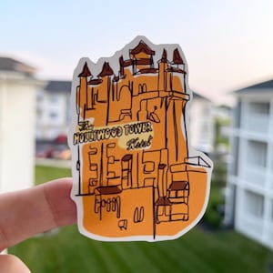 May include: A sticker featuring an orange illustration of the Hollywood Tower Hotel. The design includes the hotel's name in black text, with a stylized, vintage aesthetic. The sticker is held against a blurred outdoor background.