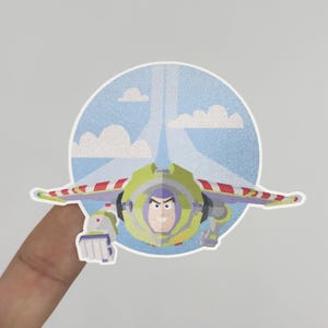May include: A colorful sticker featuring Buzz Lightyear in flight. The sticker shows Buzz with his wings extended, set against a blue sky background with clouds and a stylized trail. The design is a vibrant cartoon illustration.