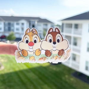 May include: A sticker featuring Chip and Dale, the Disney chipmunk duo. Chip is light brown with a black nose, and Dale is dark brown with a red nose. They are depicted peeking over a patch of grass with acorns.