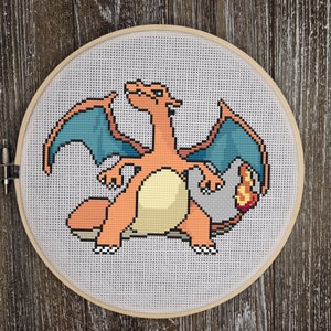 May include: A cross-stitch embroidery of a cartoon orange and blue Pokemon character with a flame tail. The character is sitting on a rock with its wings spread out.