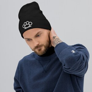 May include: A black beanie with a white knuckle duster design. The beanie is being worn by a person with a beard and a tattoo on their arm.