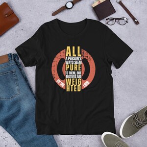 May include: Black t-shirt with a circular design in yellow and orange, featuring the text "ALL A PERSON'S WAYS SEEM PURE TO THEM, BUT MOTIVES ARE WEIGHTED BY THE LORD."