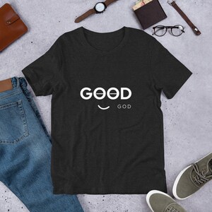 Può includere: Una maglietta grigia scura con una grafica bianca che dice "GOOD" con una faccina sorridente sotto e la parola "GOD" sotto la faccina sorridente.