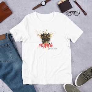 Puede incluir: Camiseta blanca con un diseño gráfico que dice "Never Stop Praying" en negro y rojo, con detalles dorados. El texto "1 Thess 5:17" está debajo. La camiseta se muestra sobre una superficie gris claro.