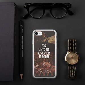 May include: Clear phone case with a religious scene and the text "FOR UNTO US A SAVIOR IS BORN." The case is next to a gold watch, black glasses, a black pencil, and a black notebook on a dark surface.