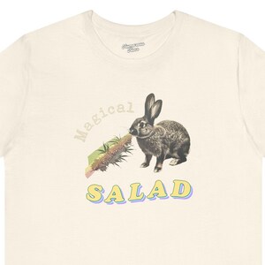 Könnte beinhalten: Ein weißes T-Shirt mit einem Aufdruck eines Kaninchens, das einen Salat isst, mit dem Text "Magical Salad" in gelben und blauen Buchstaben.