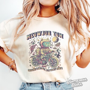 May include: A cream colored t-shirt with a graphic of a cat wearing a space helmet and riding a motorcycle. The text "MEOWJOR TOM" is above the cat and "GROUND CONTROL TO FURRY PAWS" is below the cat.