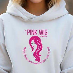 Puede incluir: Sudadera con capucha blanca con un logotipo rosa que dice "The Pink Wig Foundation" y presenta una silueta rosa de una mujer con el pelo largo. El texto alrededor del logotipo dice "Helping women with cancer in their time of need."