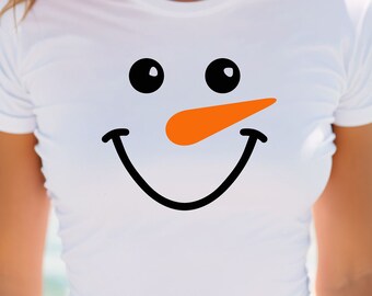 May include: White t-shirt featuring a snowman face design. The design includes black eyes, an orange carrot nose, and a black smiling mouth. The shirt is short-sleeved and appears to be made of a lightweight material.