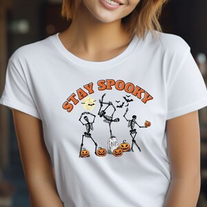 Puede incluir: Camiseta blanca con un diseño de Halloween. El diseño presenta tres esqueletos bailando, calabazas, una lápida, murciélagos y las palabras "Stay Spooky" en naranja. Una camiseta divertida y festiva.