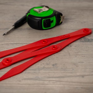 May include: A red divider tool with the word "Patio" printed on it, a green and black tape measure, and a black pencil are arranged on a wooden surface. The tools are used for measuring and marking.