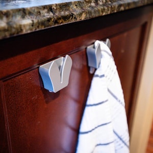 May include: Two silver-colored towel hooks mounted on a dark brown cabinet door. A white and blue striped towel hangs from one of the hooks. The hooks have a modern, minimalist design, and are likely made of metal.