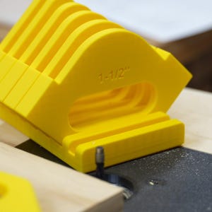 May include: Bright yellow woodworking jig with a series of curved slots. The jig is marked with "1-1/2" and is positioned on a wooden surface near a router bit. The tool appears to be designed for precise cuts.