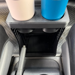 May include: A black plastic vehicle center console organizer featuring two top cup holders currently holding a light pink tumbler and a bright blue tumbler, with an open storage compartment located directly beneath the cup holder section.