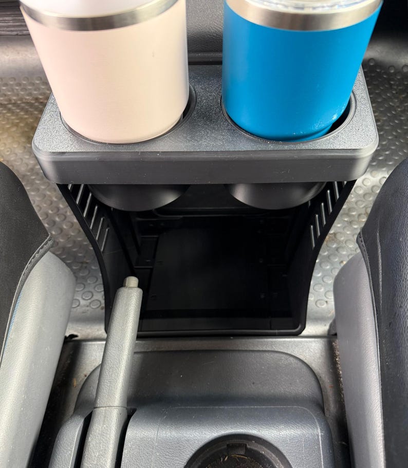 May include: A black plastic vehicle center console organizer featuring two top cup holders currently holding a light pink tumbler and a bright blue tumbler, with an open storage compartment located directly beneath the cup holder section.