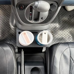 May include: A black plastic car cup holder insert positioned in the center console, featuring two circular slots holding a light pink tumbler and a dark blue tumbler, each with a white lid and a rectangular handle, situated below the gear shift.