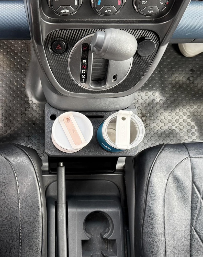 May include: A black plastic car cup holder insert positioned in the center console, featuring two circular slots holding a light pink tumbler and a dark blue tumbler, each with a white lid and a rectangular handle, situated below the gear shift.