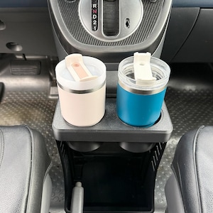May include: Two insulated tumblers with white flip-top lids sit in a black plastic cup holder insert. One tumbler is matte light pink, and the other is matte blue. The cup holder is positioned in the center console of a vehicle near the gear shifter.