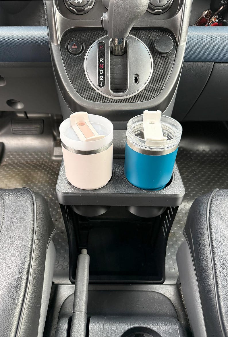 May include: Two insulated tumblers with white flip-top lids sit in a black plastic cup holder insert. One tumbler is matte light pink, and the other is matte blue. The cup holder is positioned in the center console of a vehicle near the gear shifter.