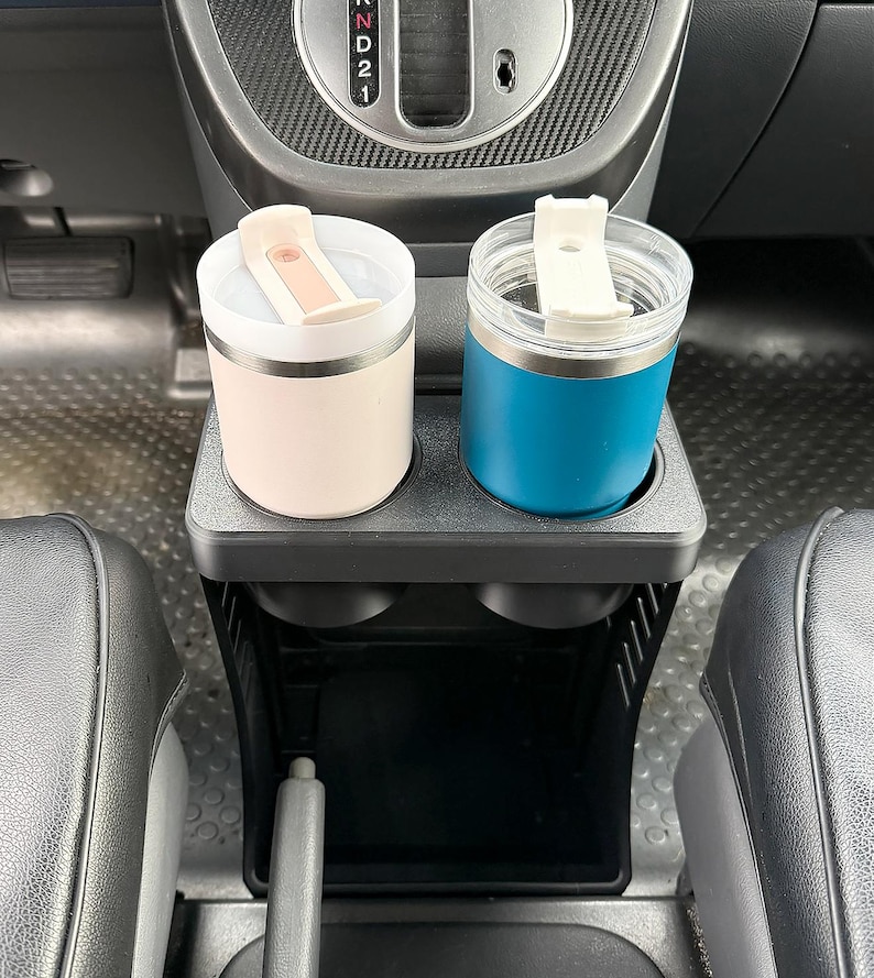 May include: Two insulated tumblers with lids sit in a black plastic car cup holder. The left tumbler is light beige with a matching lid, and the right tumbler is teal with a clear lid and a white flip-top closure. The gear shift is visible above.