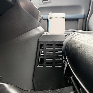 May include: A black center console organizer with a grid-like vented side panel installed between the vehicle seats. A light-colored cylindrical travel mug with a white lid sits securely in the integrated cup holder on top of the console unit.