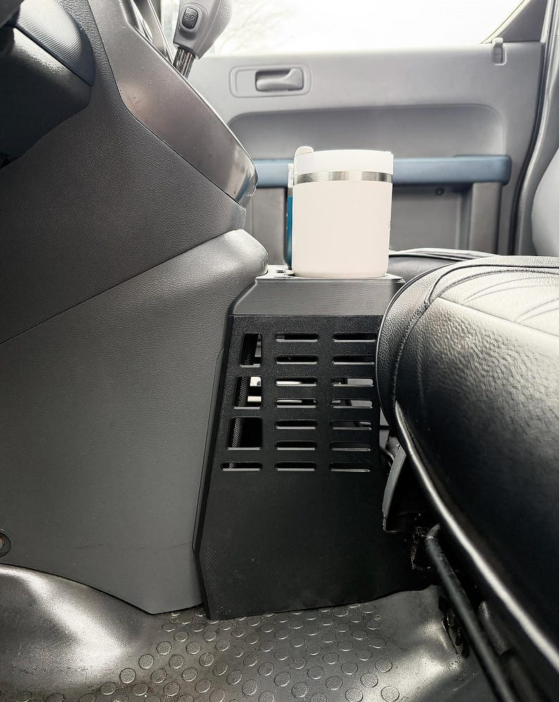 May include: A black center console organizer with a grid-like vented side panel installed between the vehicle seats. A light-colored cylindrical travel mug with a white lid sits securely in the integrated cup holder on top of the console unit.
