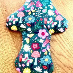 May include: A mushroom-shaped fabric item with a dark teal background. The fabric is patterned with colorful flowers, butterflies, and mushrooms. The mushrooms have red and white polka dots. The item is likely a decorative pillow or cushion.