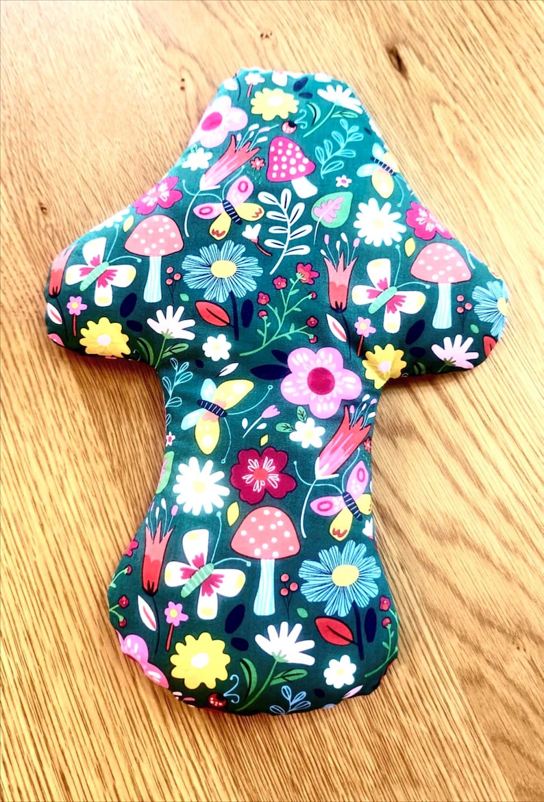 May include: A mushroom-shaped fabric item with a dark teal background. The fabric is patterned with colorful flowers, butterflies, and mushrooms. The mushrooms have red and white polka dots. The item is likely a decorative pillow or cushion.