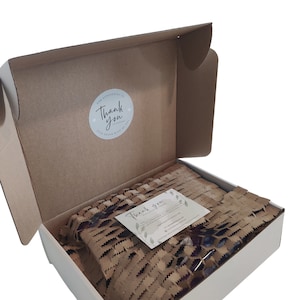 May include: An open cardboard box with a white interior, filled with brown packing paper and a small card. The box has a round sticker that says "Thank you" and the text "Your order made my day".