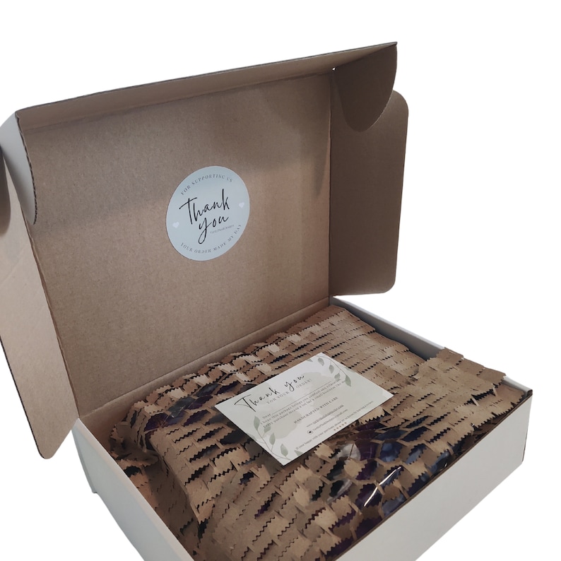 May include: An open cardboard box with a white interior, filled with brown packing paper and a small card. The box has a round sticker that says "Thank you" and the text "Your order made my day".