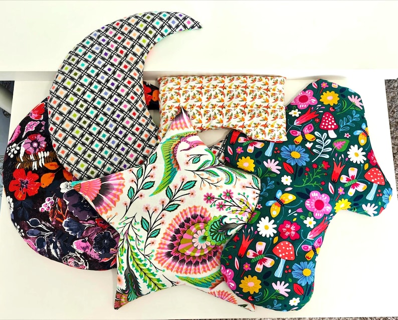 May include: A collection of colorful, patterned fabric items. Includes a crescent-shaped pillow with a black and white grid design, a star-shaped pillow with floral and leaf patterns, and a rectangular pillow with a fall-themed print.